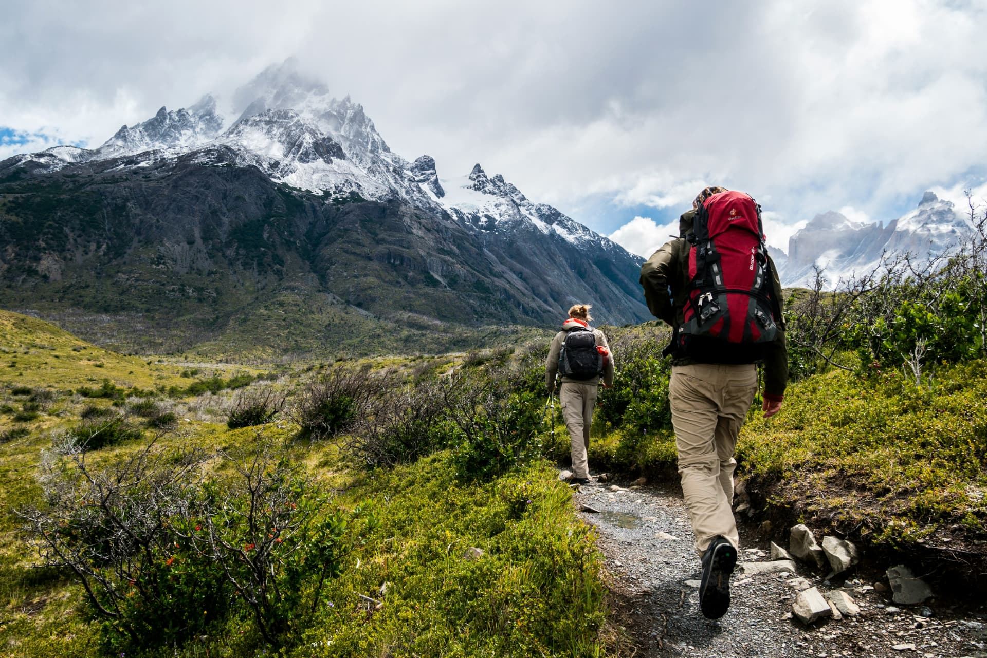 Two hikers with backpacks walking a mountain trail toward snowy, jagged peaks