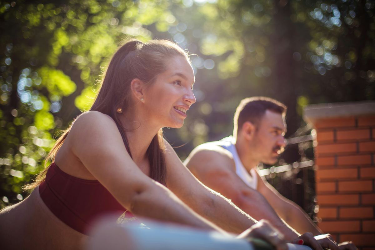 A woman and a man stretching outdoors in warm sunlight beside a brick wall
